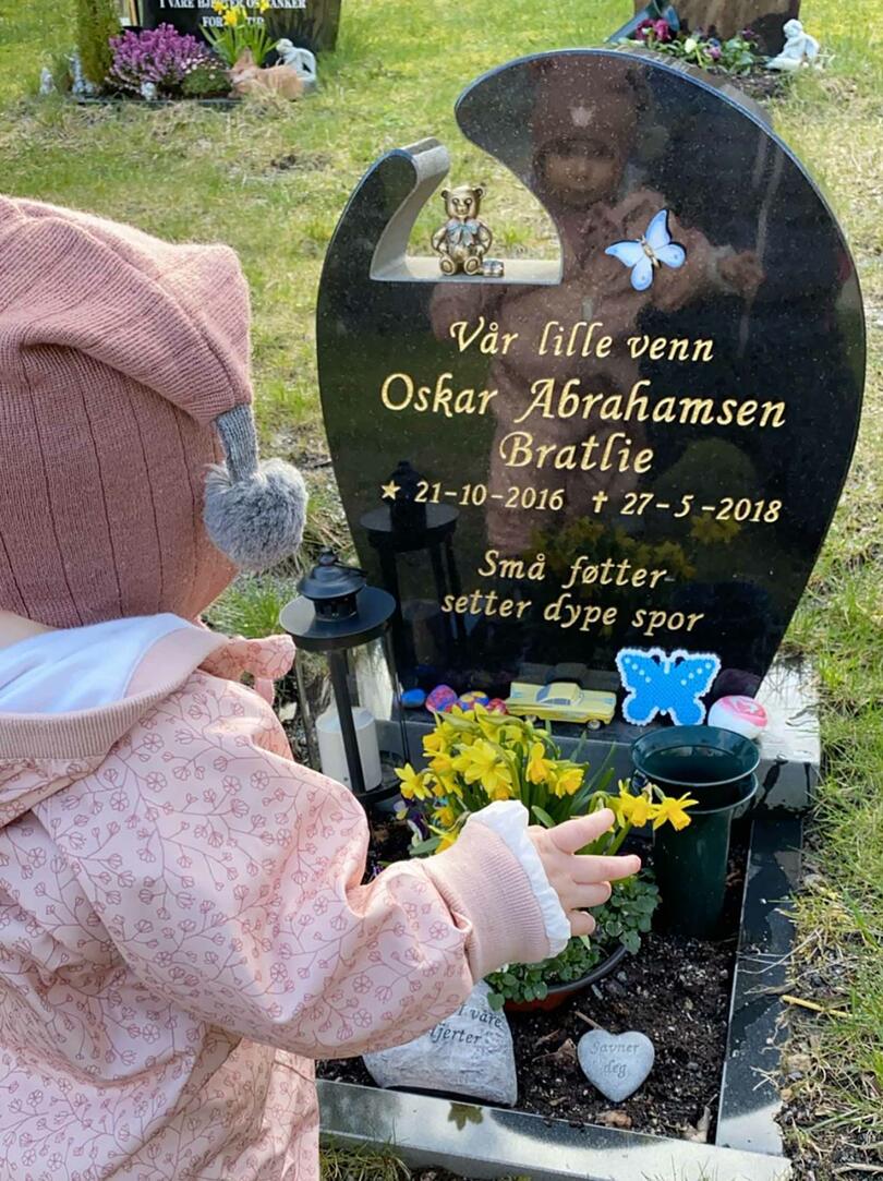 Bilde av Solveig som står foran graven til Oskar, vi ser henne bakfra, kledd i rosa lue og jakke,og kan skimte speilbiidet hennes i gravstenen.