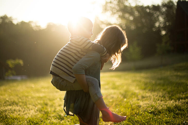 En jente bærer lillebror på ryggen, på gresslette i sollys