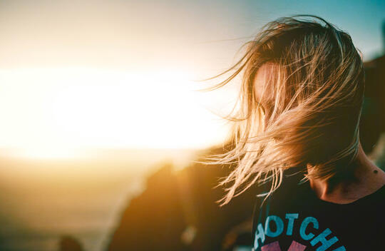 ung kvinne står ute, solnedgang i bakgrunnen, ser ned, håret blåser foran ansiktet.